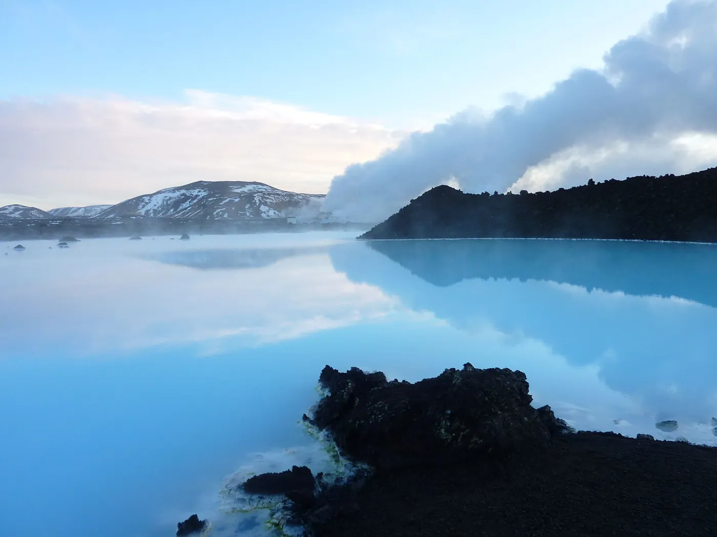 Milchig-blaues Wasser der Blauen Lagune zwischen schwarzen Lavafelsen, im Hintergrund Dampf und schneebedeckte Berge
