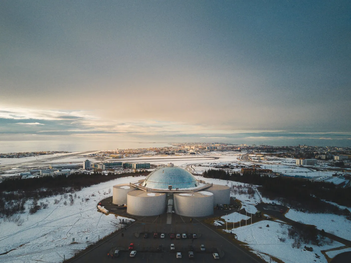 Perlan mit Glaskuppel und Warmwassertanks auf dem verschneiten Hügel Öskjuhlíð, dahinter Reykjavík und das Meer (Luftbild)