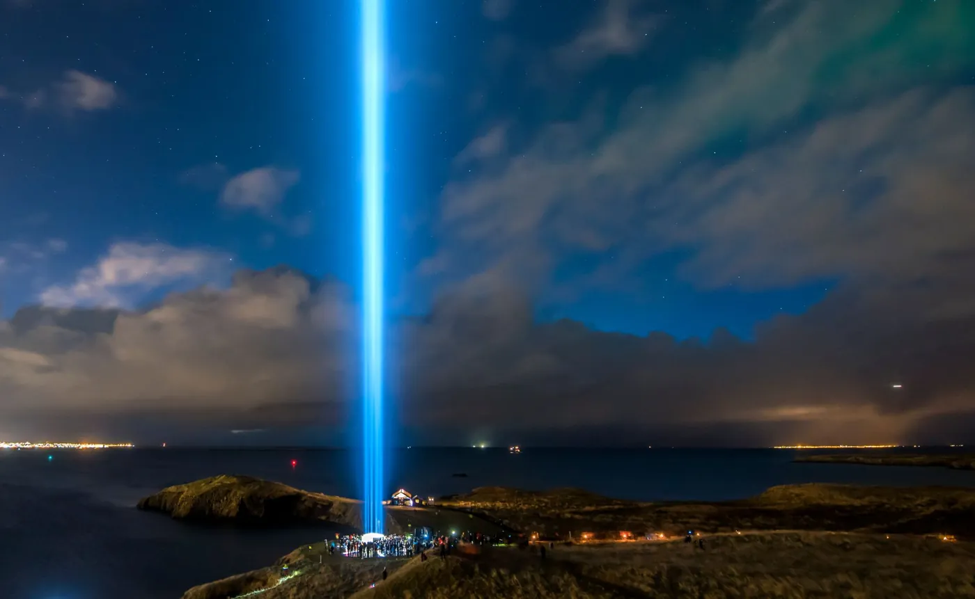 Der Lichtstrahl des Imagine Peace Tower auf der Insel Viðey bei Nacht, im Hintergrund die Lichter Reykjavíks